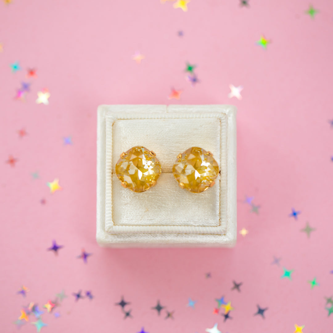 A pair of mustard yellow faceted cushion cut stud earrings displayed on a cream-colored jewelry box against a pink background with scattered star-shaped confetti.