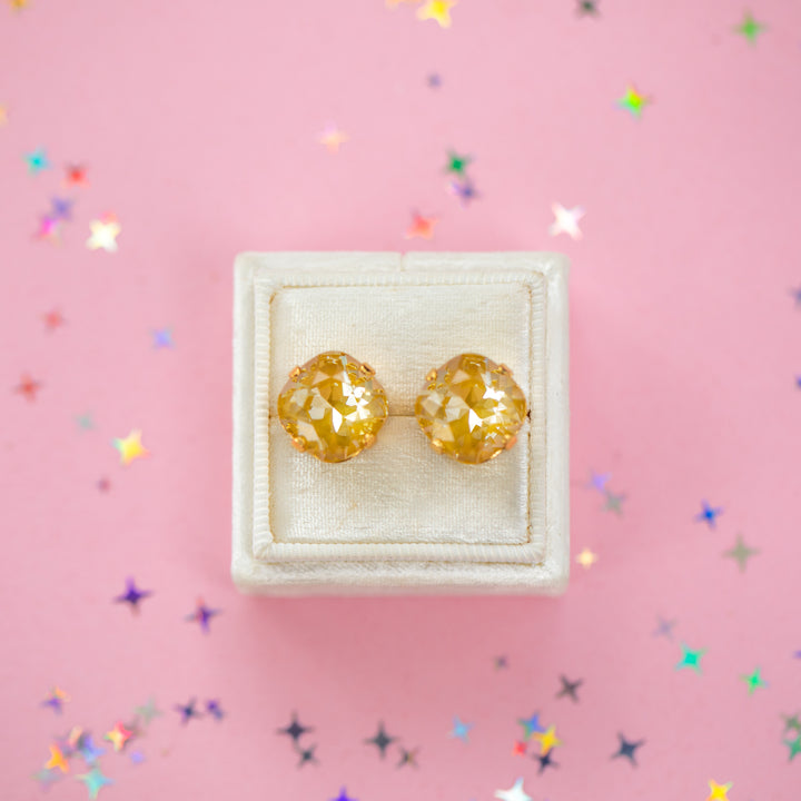 A pair of mustard yellow faceted cushion cut stud earrings displayed on a cream-colored jewelry box against a pink background with scattered star-shaped confetti.