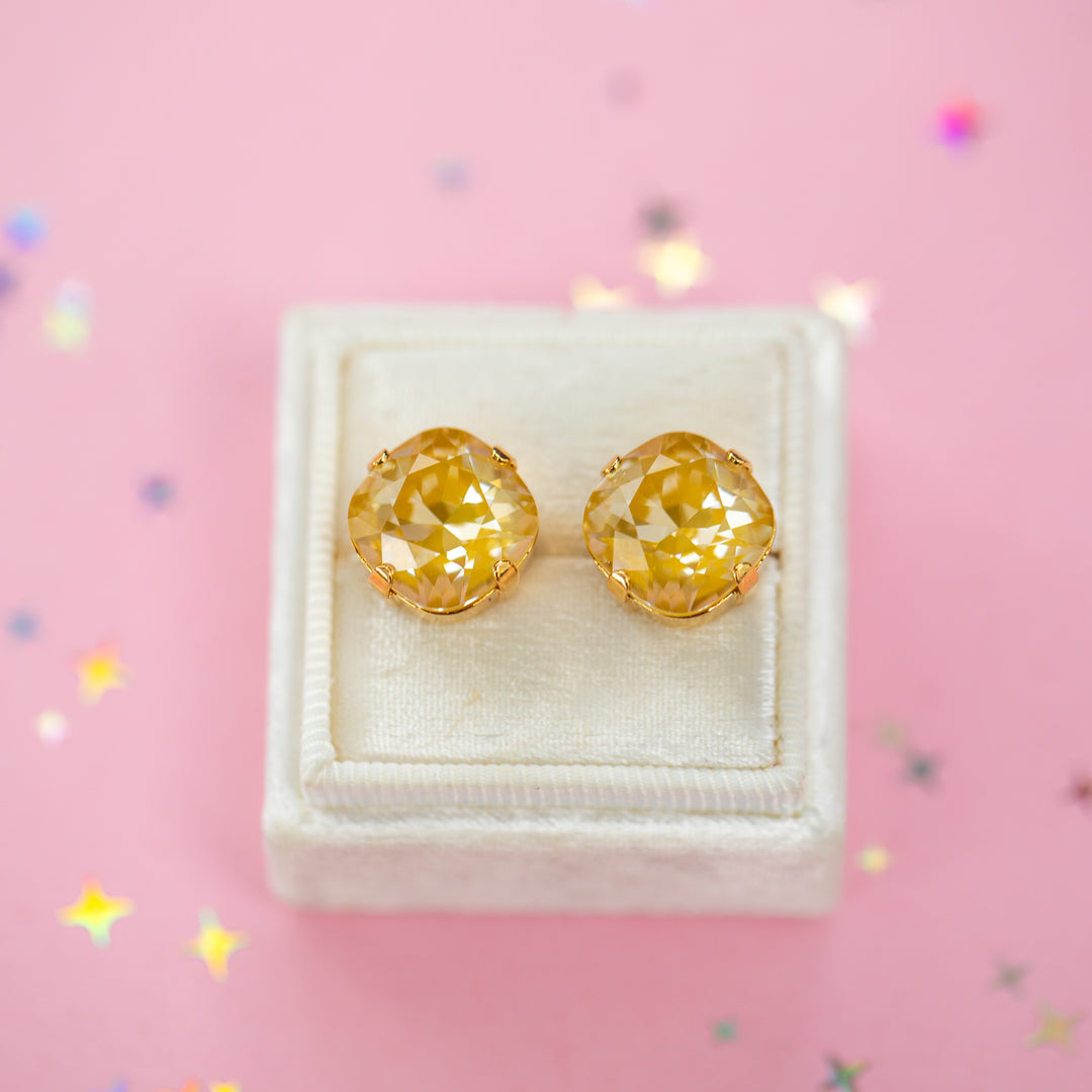 A pair of mustard yellow faceted cushion cut stud earrings displayed on a cream-colored jewelry box against a pink background with scattered star-shaped confetti.