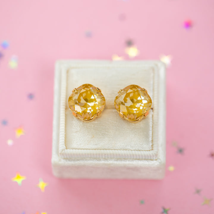 A pair of mustard yellow faceted cushion cut stud earrings displayed on a cream-colored jewelry box against a pink background with scattered star-shaped confetti.
