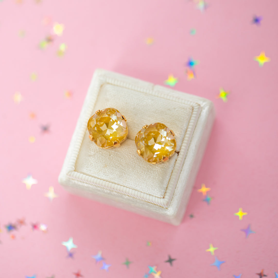 A pair of mustard yellow faceted cushion cut stud earrings displayed on a cream-colored jewelry box against a pink background with scattered star-shaped confetti.