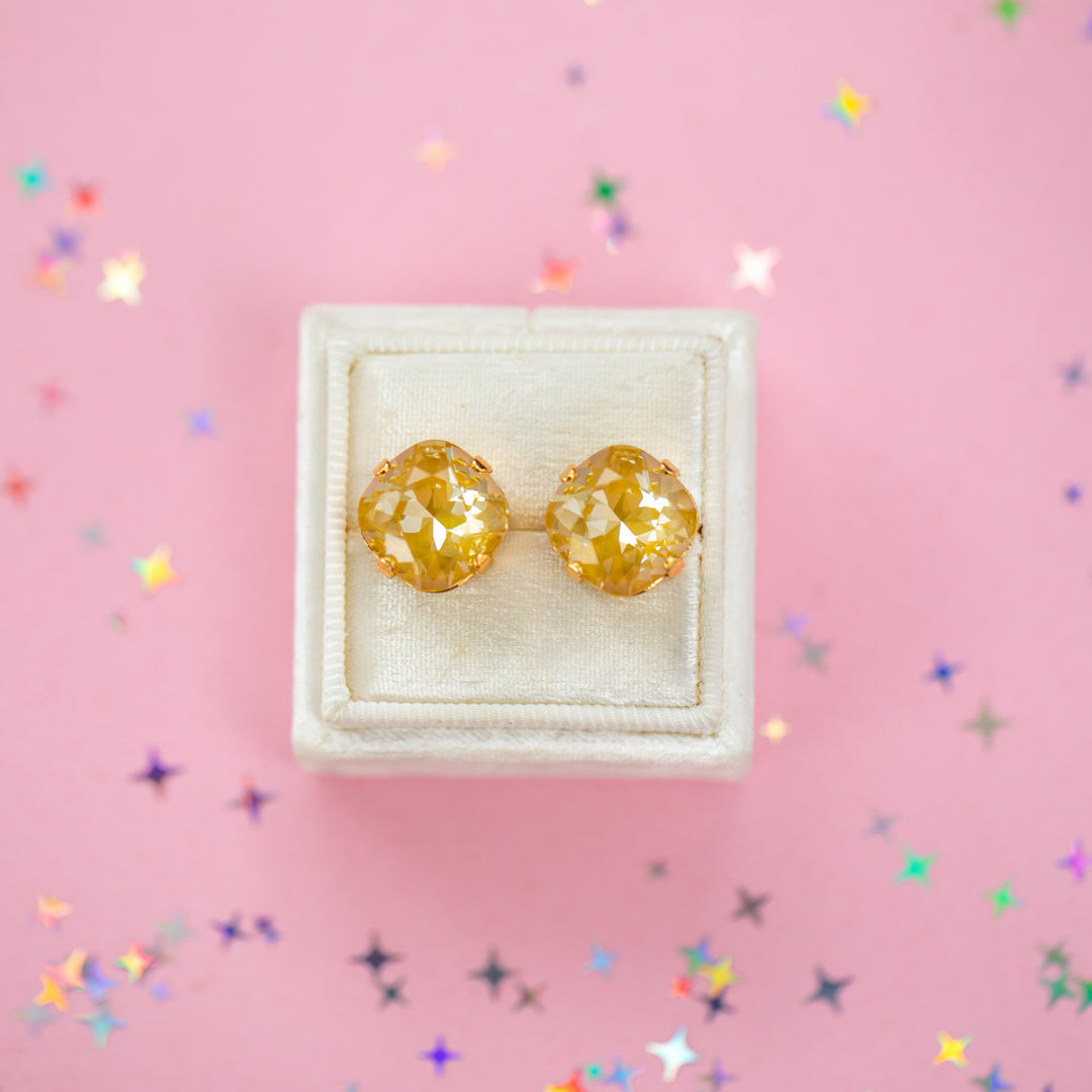 A pair of mustard yellow faceted cushion cut stud earrings displayed on a cream-colored jewelry box against a pink background with scattered star-shaped confetti.