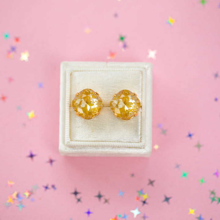 A pair of mustard yellow faceted cushion cut stud earrings displayed on a cream-colored jewelry box against a pink background with scattered star-shaped confetti.