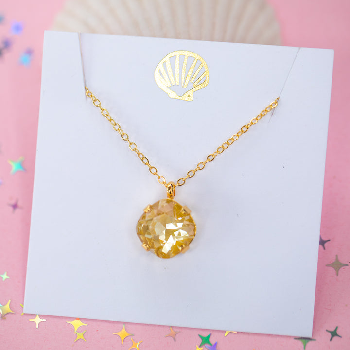 A gold chain necklace with a cushion cut faceted yellow crystal pendant, displayed on a white card with a gold seashell design, set against a pink background with star confetti.