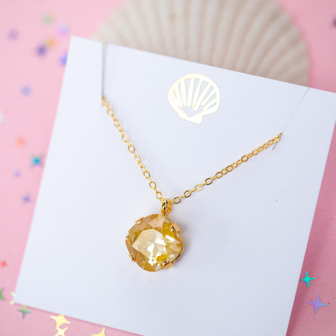 A gold chain necklace with a cushion cut faceted yellow crystal pendant, displayed on a white card with a gold seashell design, set against a pink background with star confetti.