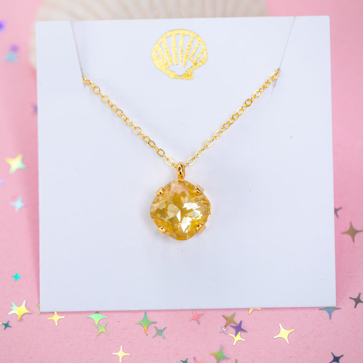 A gold chain necklace with a cushion cut faceted yellow crystal pendant, displayed on a white card with a gold seashell design, set against a pink background with star confetti.