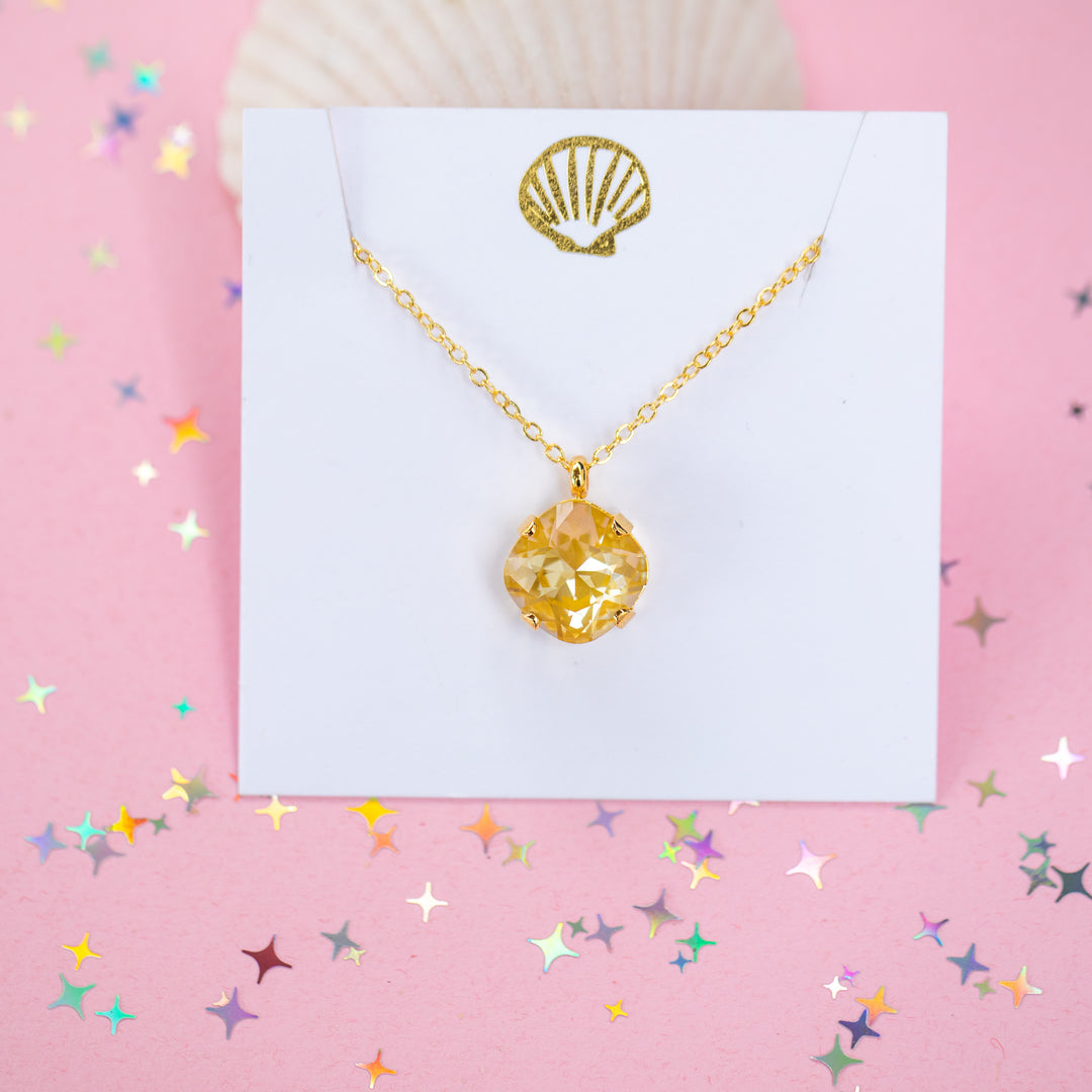 A gold chain necklace with a cushion cut faceted yellow crystal pendant, displayed on a white card with a gold seashell design, set against a pink background with star confetti.