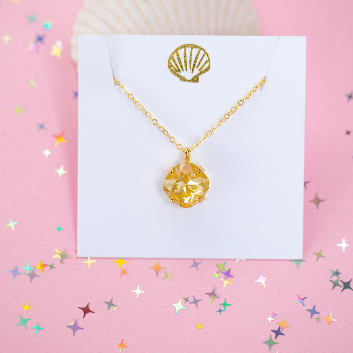 A gold chain necklace with a cushion cut faceted yellow crystal pendant, displayed on a white card with a gold seashell design, set against a pink background with star confetti.