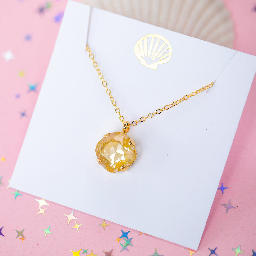 A gold chain necklace with a cushion cut faceted yellow crystal pendant, displayed on a white card with a gold seashell design, set against a pink background with star confetti.