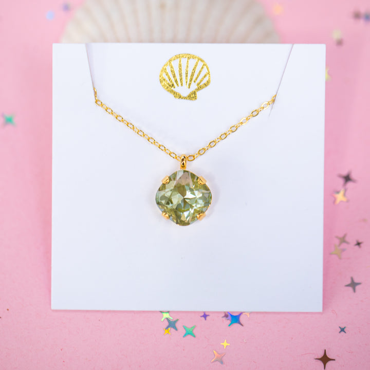 A gold chain necklace with a faceted cushion cut olive green crystal pendant, displayed on a white card with a small gold seashell illustration, set against a pink background with scattered star confetti.