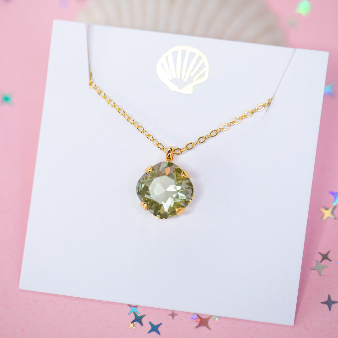 A gold chain necklace with a faceted cushion cut olive green crystal pendant, displayed on a white card with a small gold seashell illustration, set against a pink background with scattered star confetti.