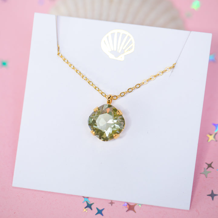 A gold chain necklace with a faceted cushion cut olive green crystal pendant, displayed on a white card with a small gold seashell illustration, set against a pink background with scattered star confetti.