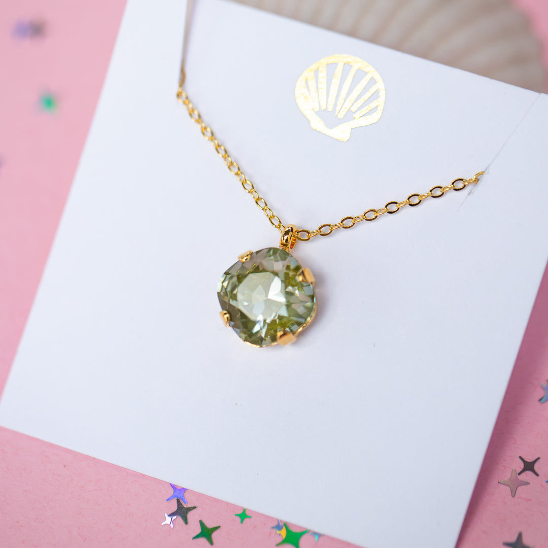 A gold chain necklace with a faceted cushion cut olive green crystal pendant, displayed on a white card with a small gold seashell illustration, set against a pink background with scattered star confetti.