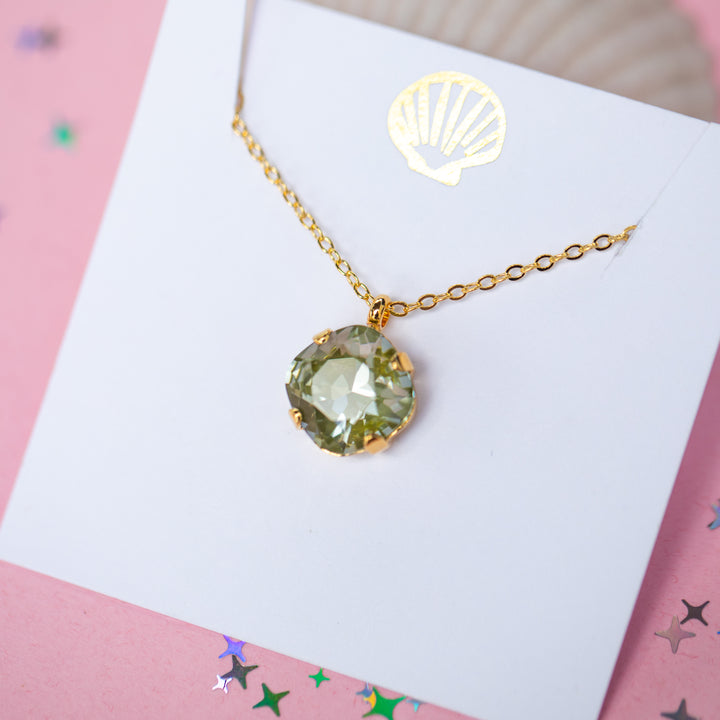 A gold chain necklace with a faceted cushion cut olive green crystal pendant, displayed on a white card with a small gold seashell illustration, set against a pink background with scattered star confetti.