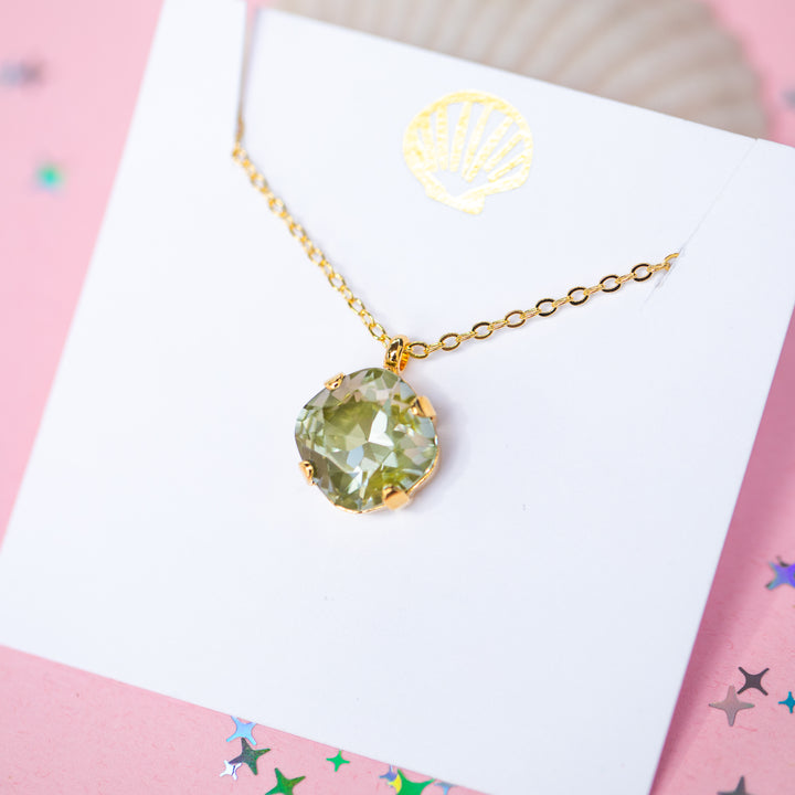 A gold chain necklace with a faceted cushion cut olive green crystal pendant, displayed on a white card with a small gold seashell illustration, set against a pink background with scattered star confetti.