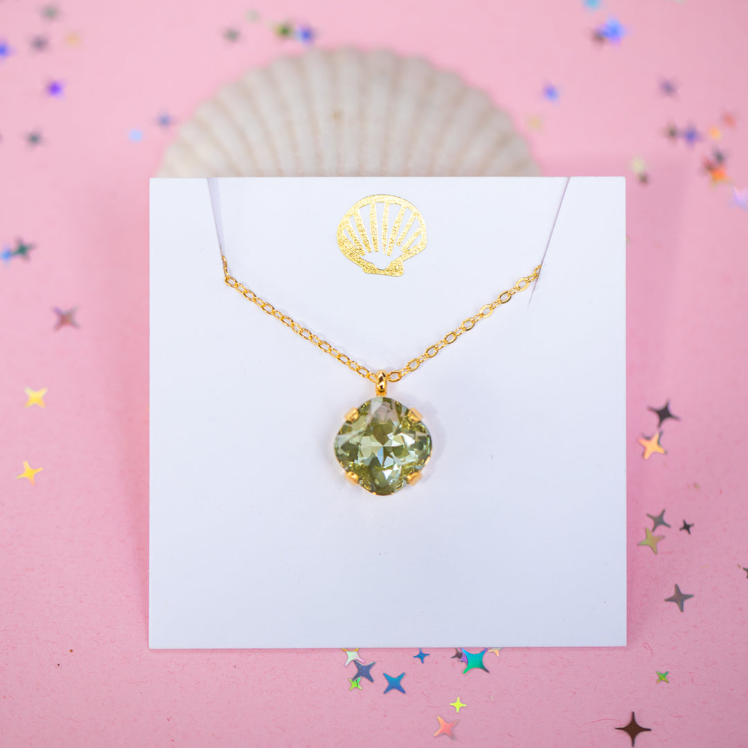 A gold chain necklace with a faceted cushion cut olive green crystal pendant, displayed on a white card with a small gold seashell illustration, set against a pink background with scattered star confetti.