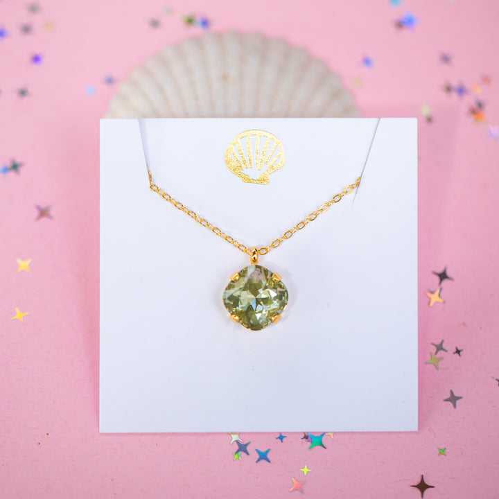 A gold chain necklace with a faceted cushion cut olive green crystal pendant, displayed on a white card with a small gold seashell illustration, set against a pink background with scattered star confetti.