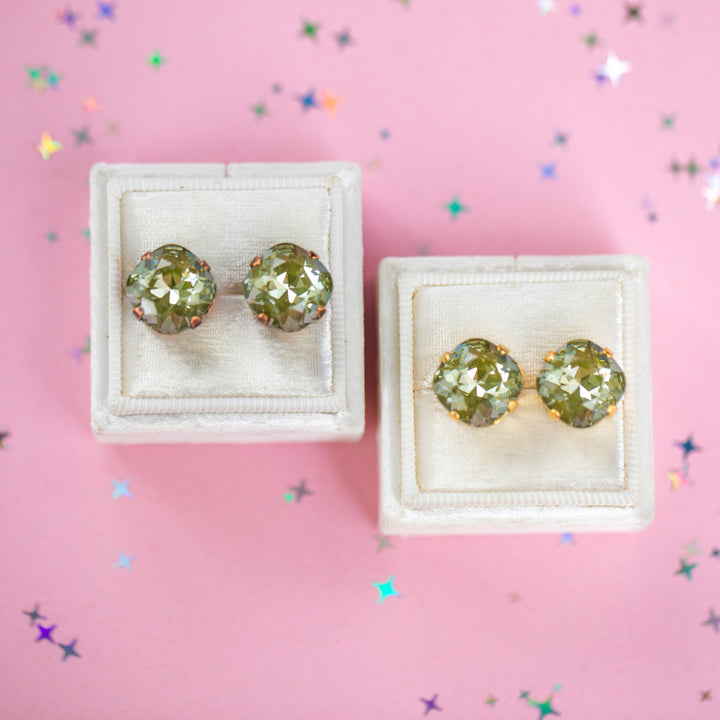 A pair of Cushion cut stud earrings with faceted olive green stones in a gold and antique copper setting, displayed on two small white jewelry boxes against a pink background with scattered iridescent star confetti.