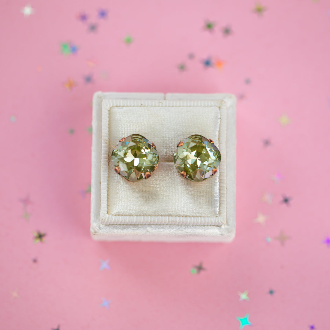 A pair of cushion-cut olive green gemstone stud earrings in antique copper settings, presented on a cream-colored velvet jewelry box against a pink background with scattered star confetti.