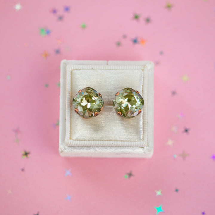 A pair of cushion-cut olive green gemstone stud earrings in antique copper settings, presented on a cream-colored velvet jewelry box against a pink background with scattered star confetti.