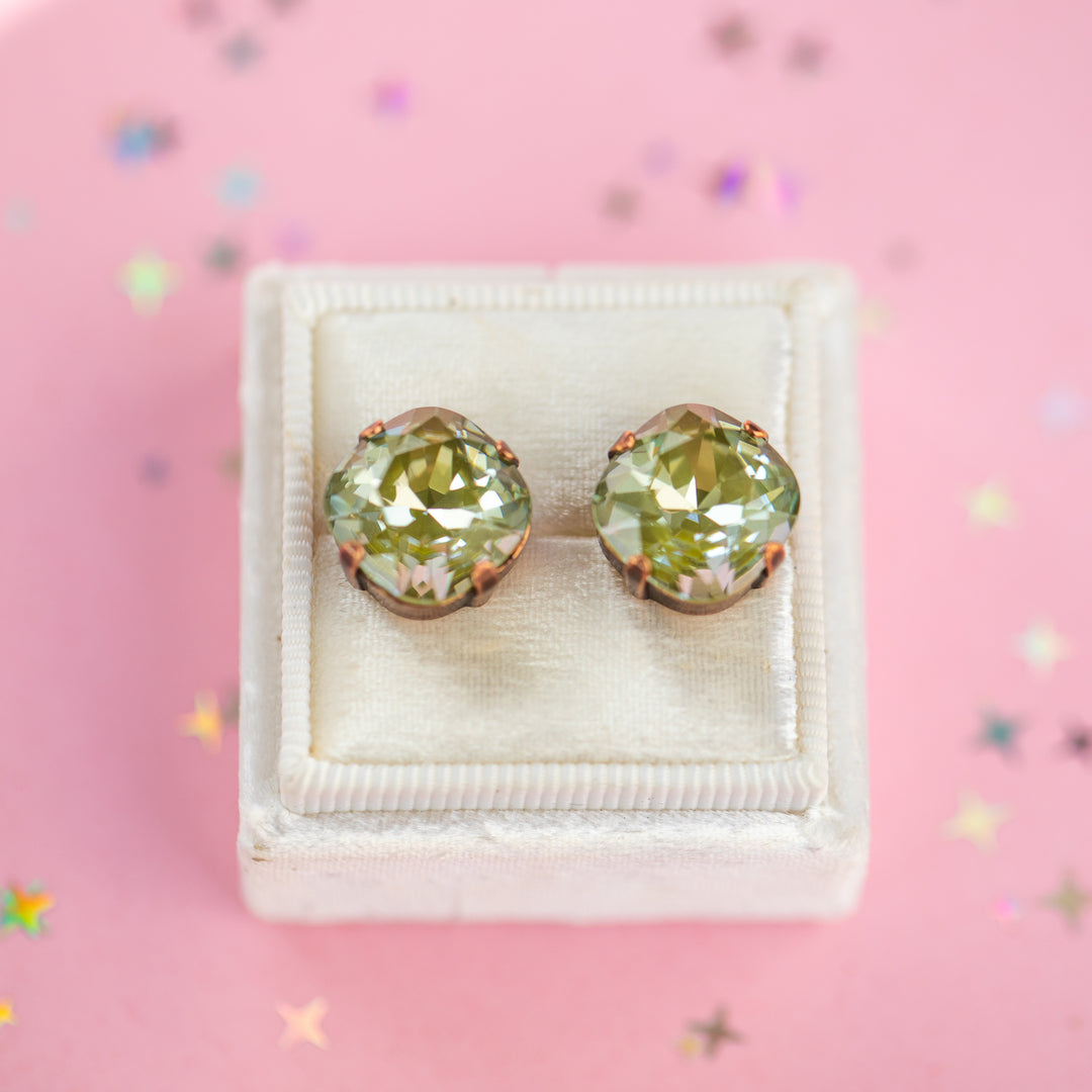 A pair of cushion-cut olive green gemstone stud earrings in Antique Copper settings, presented on a cream-colored velvet jewelry box against a pink background with scattered star confetti.