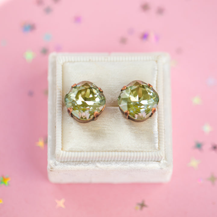 A pair of cushion-cut olive green gemstone stud earrings in Antique Copper settings, presented on a cream-colored velvet jewelry box against a pink background with scattered star confetti.