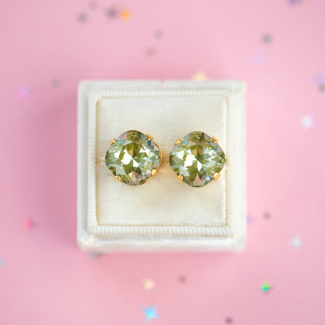 A pair of cushion-cut olive green gemstone stud earrings in gold settings, presented on a cream-colored velvet jewelry box against a pink background with scattered star confetti.