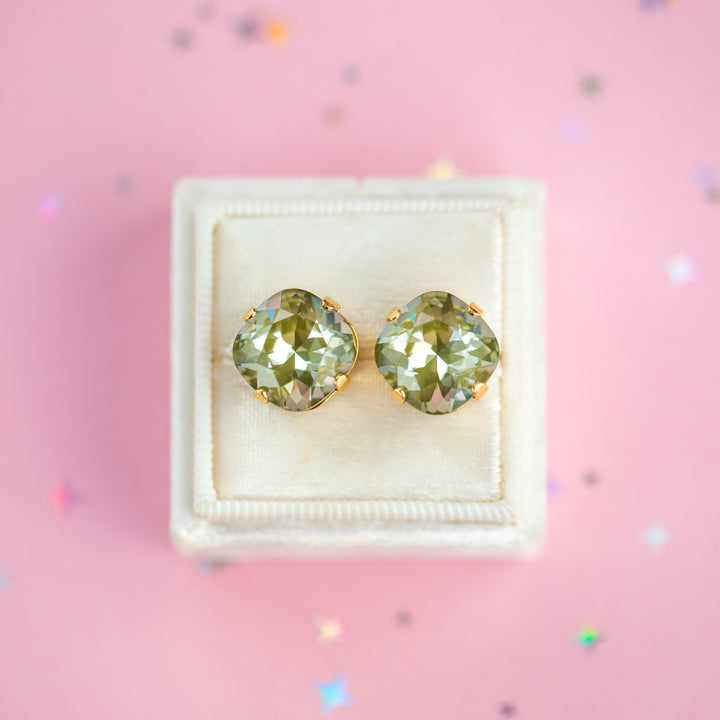 A pair of cushion-cut olive green gemstone stud earrings in gold settings, presented on a cream-colored velvet jewelry box against a pink background with scattered star confetti.