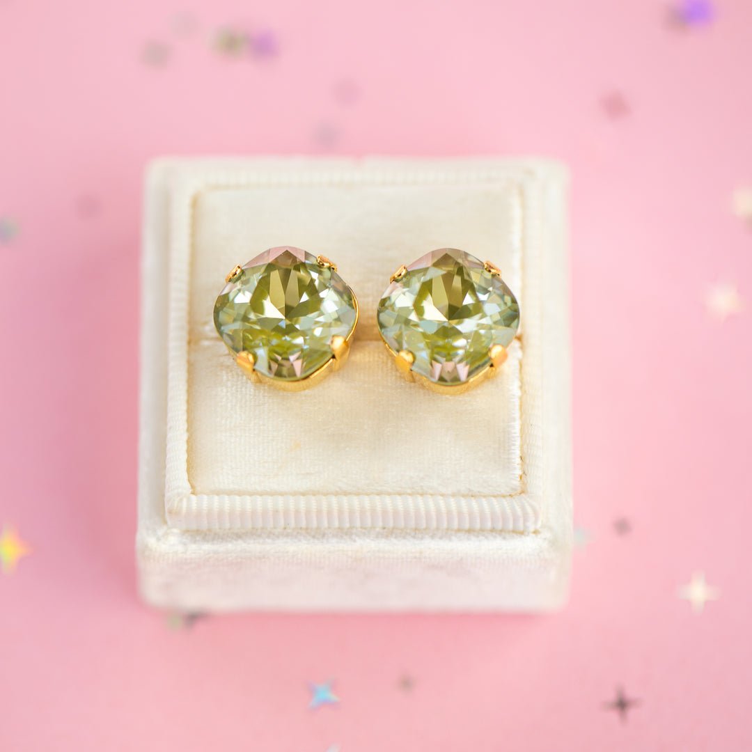 A pair of cushion-cut olive green gemstone stud earrings in gold settings, presented on a cream-colored velvet jewelry box against a pink background with scattered star confetti.