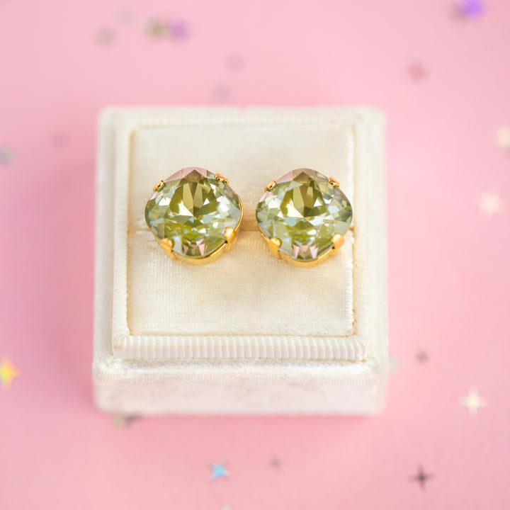 A pair of cushion-cut olive green gemstone stud earrings in gold settings, presented on a cream-colored velvet jewelry box against a pink background with scattered star confetti.