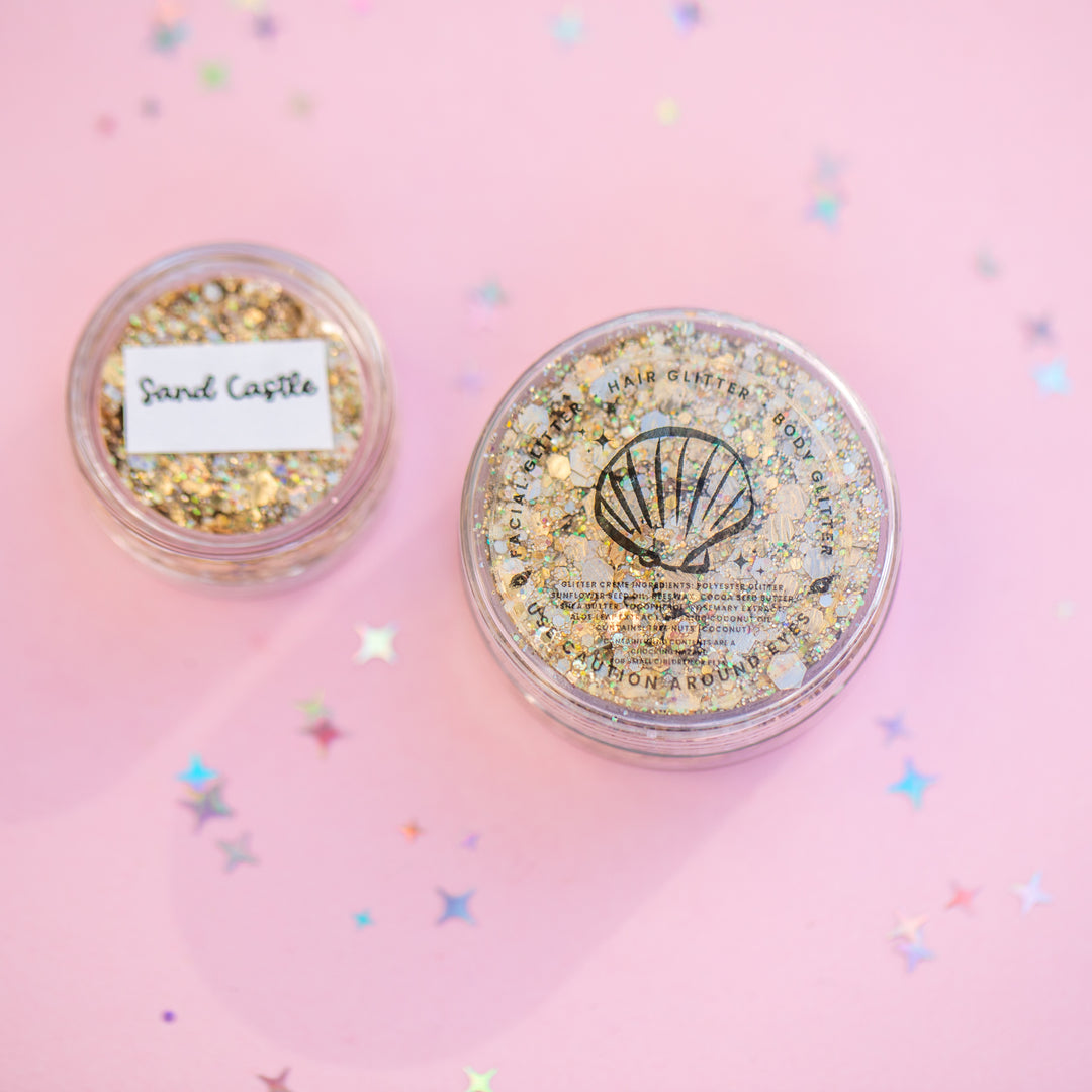 Two containers of glitter with 'Sand Castle' and 'Seashell' labels on a pink background.
