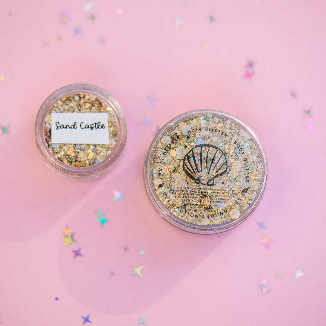 Two containers of glitter with 'Sand Castle' and 'Shell' labels on a pink background with confetti.