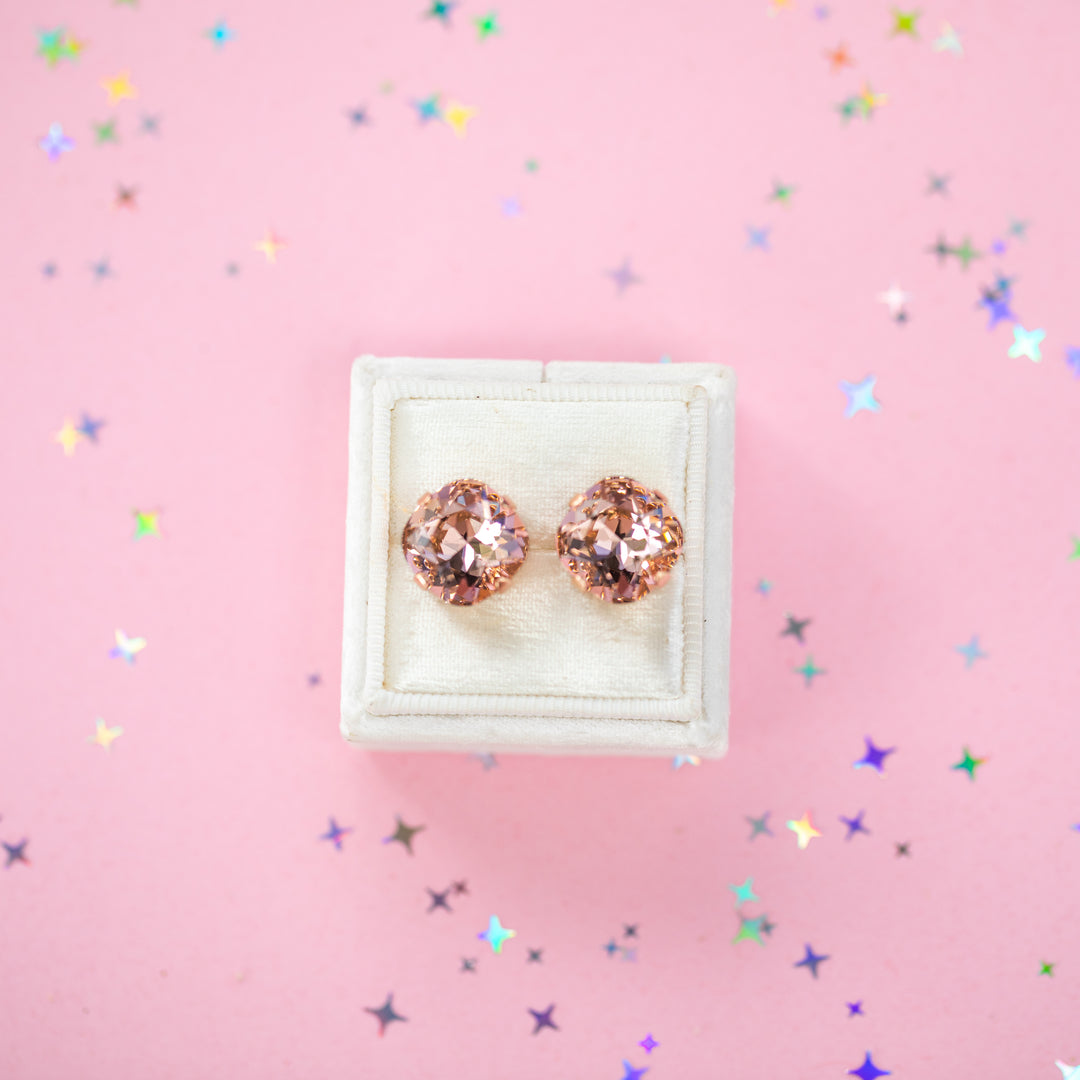 A pair of rose gold stud earrings with cushion cut faceted warm toned pink gemstone crystals, displayed on a cream-colored jewelry box against a soft pink background with subtle star confetti.