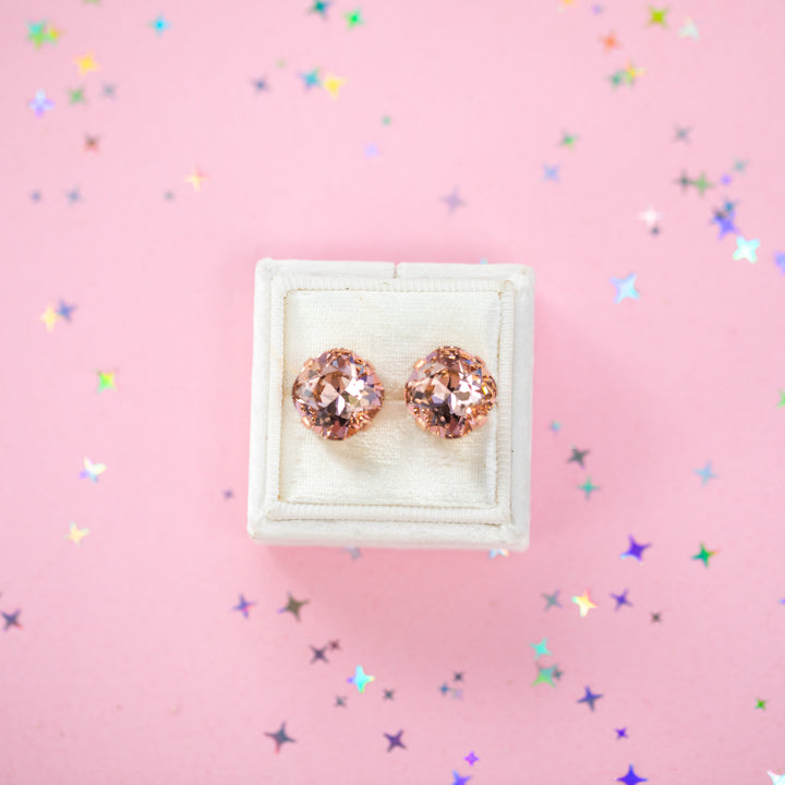 A pair of rose gold stud earrings with cushion cut faceted warm toned pink gemstone crystals, displayed on a cream-colored jewelry box against a soft pink background with subtle star confetti.