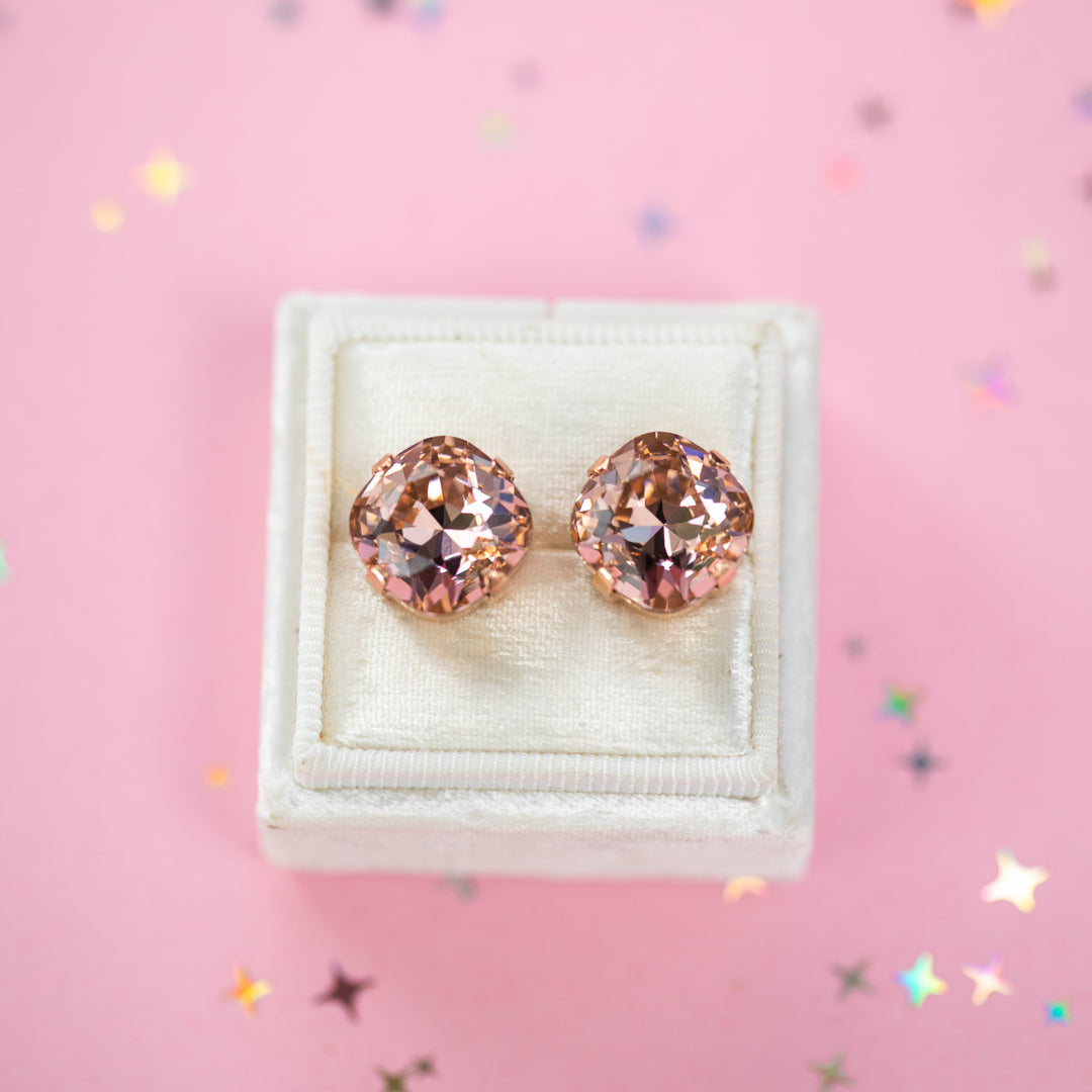 A pair of rose gold stud earrings with cushion cut faceted warm toned pink gemstone crystals, displayed on a cream-colored jewelry box against a soft pink background with subtle star confetti.