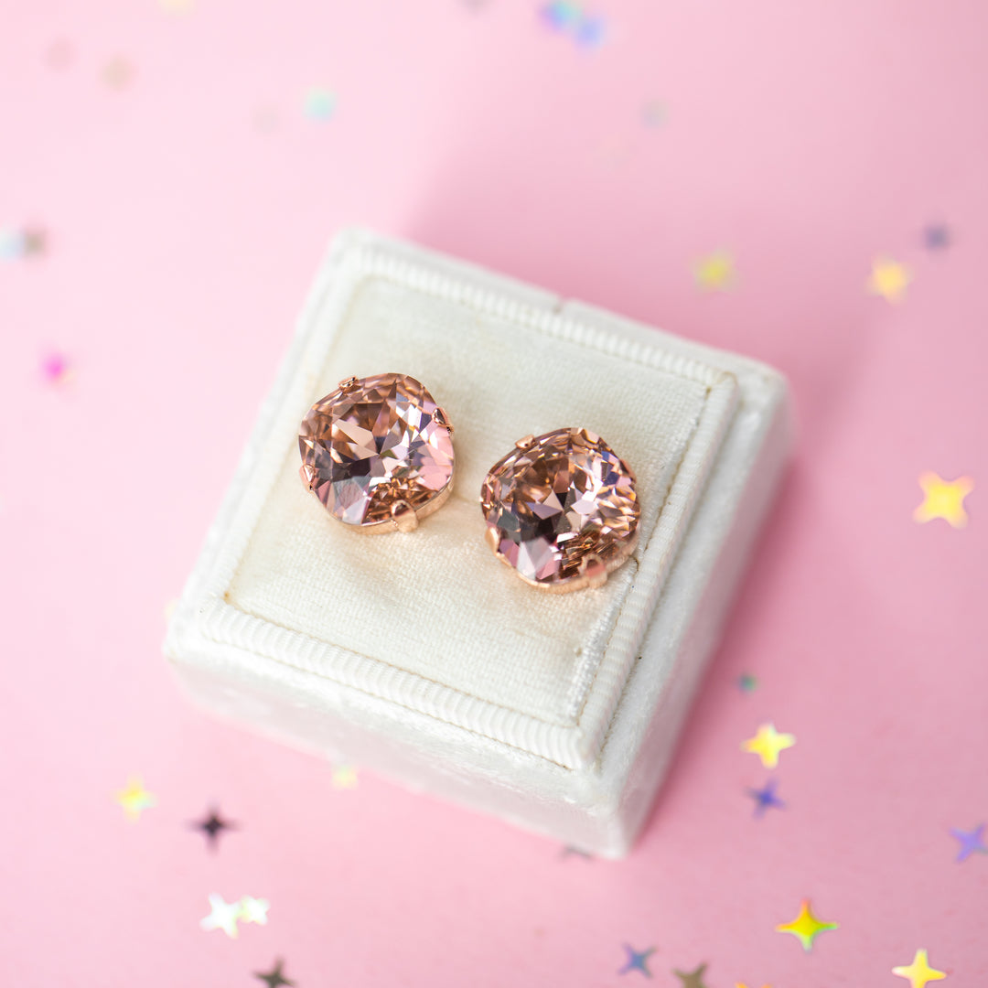 A pair of rose gold stud earrings with cushion cut faceted warm toned pink gemstone crystals, displayed on a cream-colored jewelry box against a soft pink background with subtle star confetti.