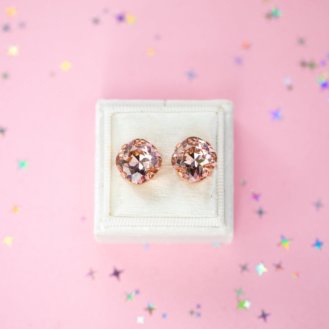 A pair of rose gold stud earrings with cushion cut faceted warm toned pink gemstone crystals, displayed on a cream-colored jewelry box against a soft pink background with subtle star confetti.