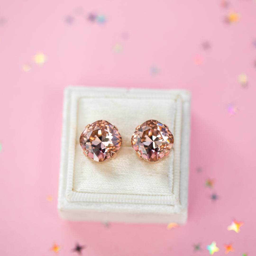 A pair of rose gold stud earrings with cushion cut faceted warm toned pink gemstone crystals, displayed on a cream-colored jewelry box against a soft pink background with subtle star confetti.