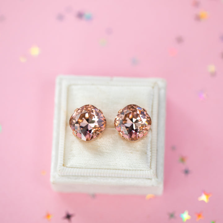 A pair of rose gold stud earrings with cushion cut faceted warm toned pink gemstone crystals, displayed on a cream-colored jewelry box against a soft pink background with subtle star confetti.