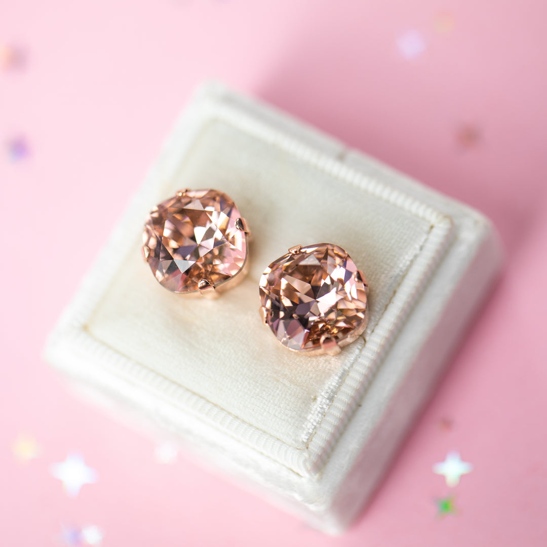 A pair of rose gold stud earrings with cushion cut faceted warm toned pink gemstone crystals, displayed on a cream-colored jewelry box against a soft pink background with subtle star confetti.