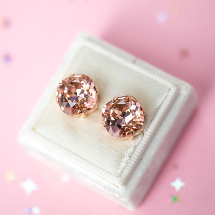A pair of rose gold stud earrings with cushion cut faceted warm toned pink gemstone crystals, displayed on a cream-colored jewelry box against a soft pink background with subtle star confetti.