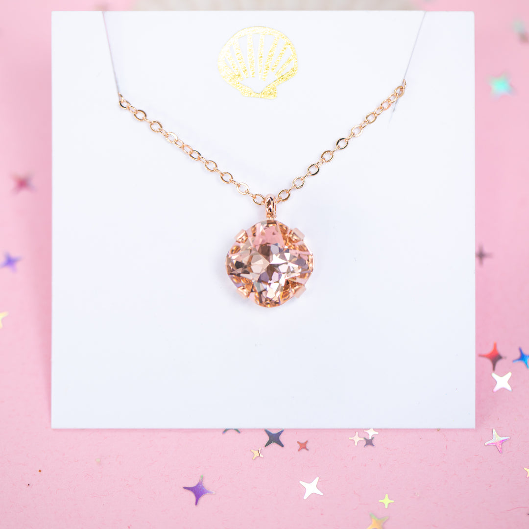 A rose gold chain necklace with a cushion cut faceted warm toned pink crystal pendant, displayed on a white card with a gold seashell logo, set against a pink background with star confetti.
