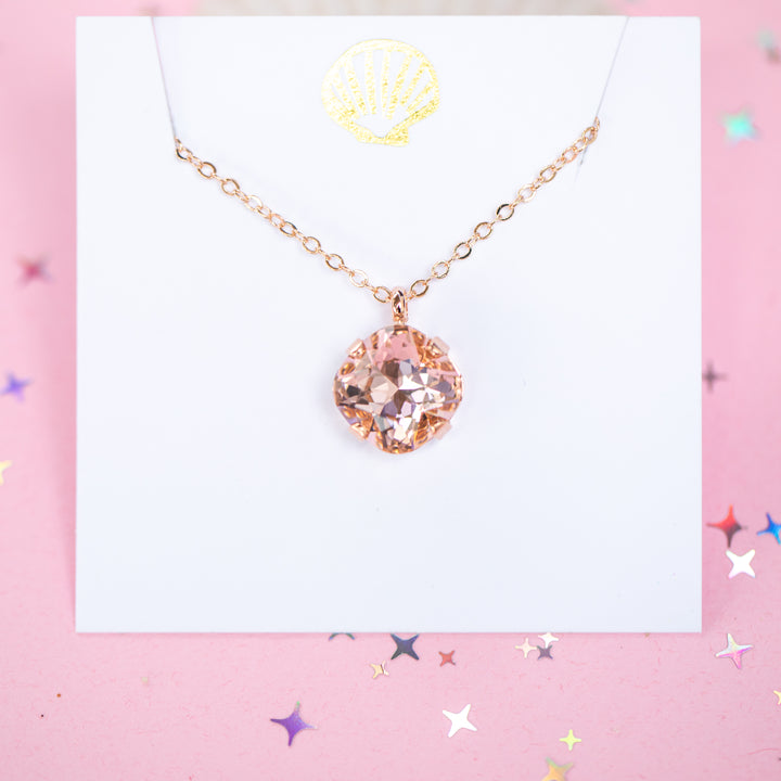 A rose gold chain necklace with a cushion cut faceted warm toned pink crystal pendant, displayed on a white card with a gold seashell logo, set against a pink background with star confetti.
