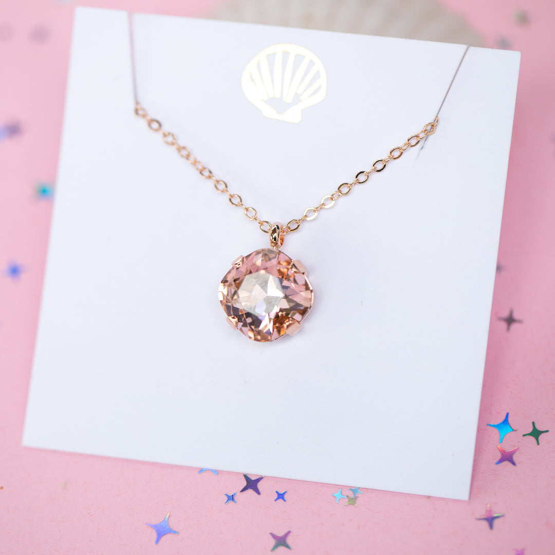 A rose gold chain necklace with a cushion cut faceted warm toned pink crystal pendant, displayed on a white card with a gold seashell logo, set against a pink background with star confetti.
