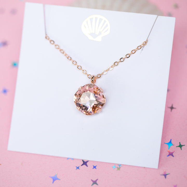 A rose gold chain necklace with a cushion cut faceted warm toned pink crystal pendant, displayed on a white card with a gold seashell logo, set against a pink background with star confetti.