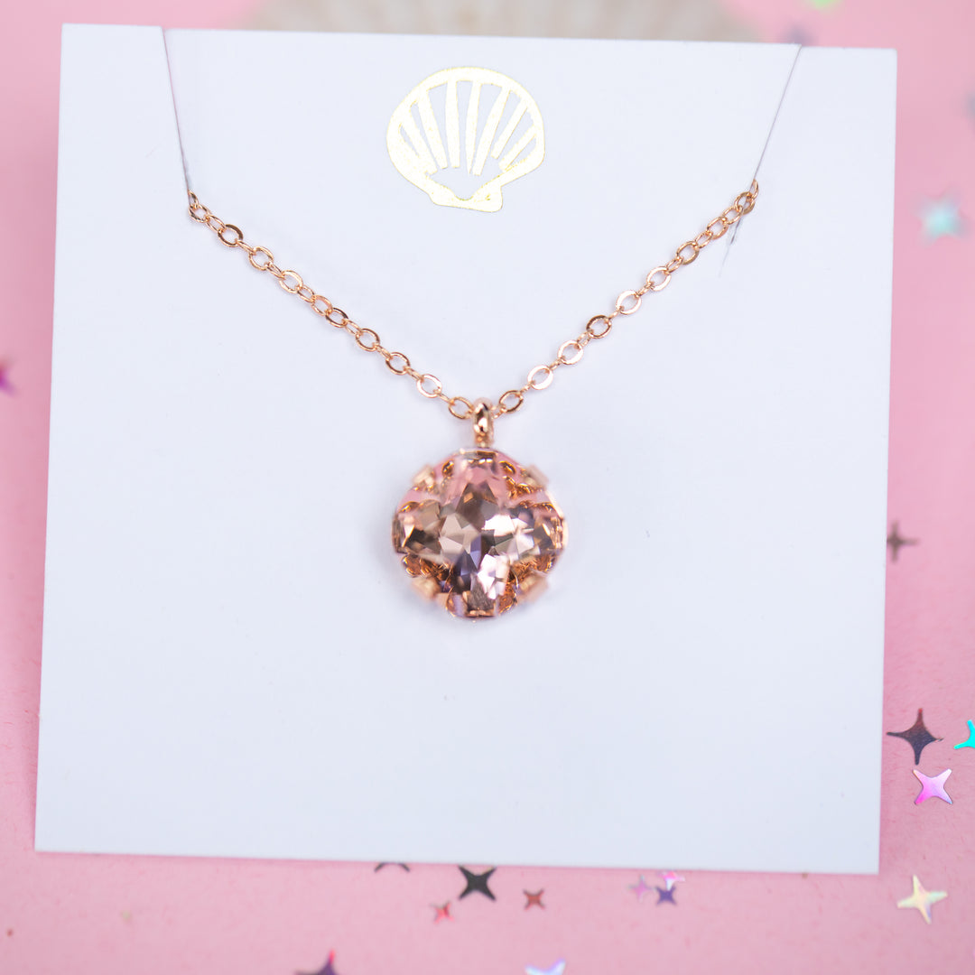 A rose gold chain necklace with a cushion cut faceted warm toned pink crystal pendant, displayed on a white card with a gold seashell logo, set against a pink background with star confetti.