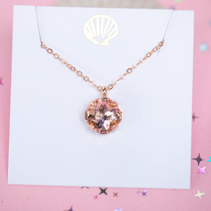 A rose gold chain necklace with a cushion cut faceted warm toned pink crystal pendant, displayed on a white card with a gold seashell logo, set against a pink background with star confetti.
