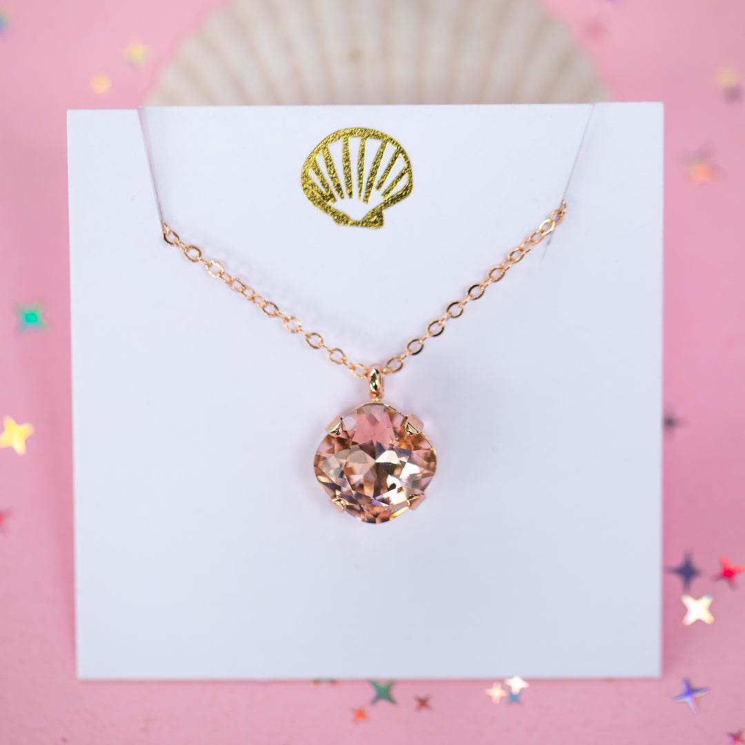 A rose gold chain necklace with a cushion cut faceted warm toned pink crystal pendant, displayed on a white card with a gold seashell logo, set against a pink background with star confetti.