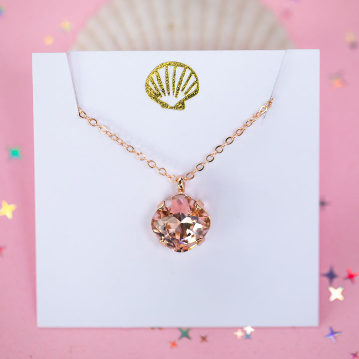 A rose gold chain necklace with a cushion cut faceted warm toned pink crystal pendant, displayed on a white card with a gold seashell logo, set against a pink background with star confetti.
