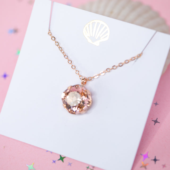 A rose gold chain necklace with a cushion cut faceted warm toned pink crystal pendant, displayed on a white card with a gold seashell logo, set against a pink background with star confetti.